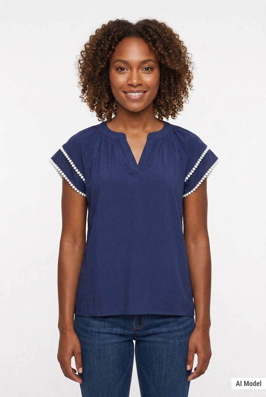 Woman wearing a navy blue gathered split-neck blouse with white pom-pom trim on flutter sleeves, paired with dark jeans,
