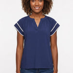 Woman wearing a navy blue gathered split-neck blouse with white pom-pom trim on flutter sleeves, paired with dark jeans,