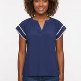 Woman wearing a navy blue gathered split-neck blouse with white pom-pom trim on flutter sleeves, paired with dark jeans,