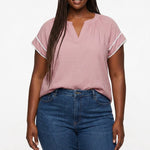 Plus-size model wears a dusty pink V-neck popover blouse with white lace-trimmed flutter sleeves, paired with blue jeans,