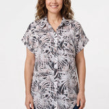 Woman wearing a short rolled-sleeve button-front blouse in black, white, and pink palm leaf print, with a collar and chest