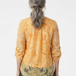 Back view of a woman with gray hair wearing an orange floral burnout blouse with rolled 3/4 sleeves and olive border hem,