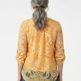 Back view of a woman with gray hair wearing an orange floral burnout blouse with rolled 3/4 sleeves and olive border hem,