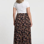 Plus-size maxi skirt in black with earth-tone floral print, back view, worn by a blonde woman with a white short-sleeve tee.