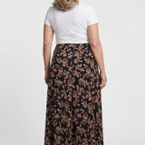 Plus-size maxi skirt in black with earth-tone floral print, back view, worn by a blonde woman with a white short-sleeve tee.