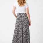 Red-haired female model shown from behind wearing a black medallion-print knit maxi skirt with a white tee and nude heels.