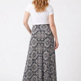 Red-haired female model shown from behind wearing a black medallion-print knit maxi skirt with a white tee and nude heels.