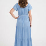 Woman wearing a light blue short-sleeve tiered maxi dress with a tie waist belt, shown from the back; white sneakers visible.