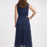 Back view of a woman in a navy sleeveless swiss dot tiered maxi dress with smocked waist, paired with nude heels.