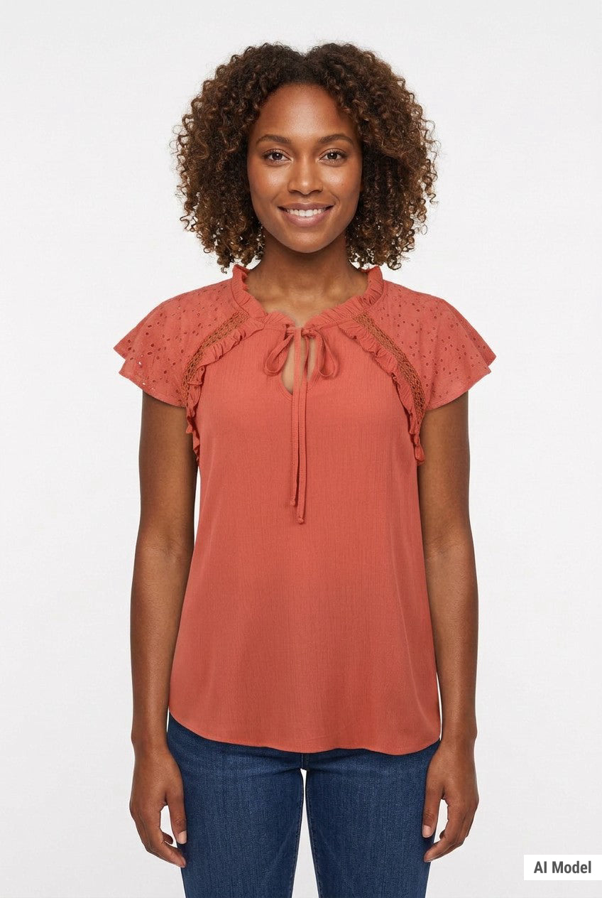 Woman wearing a rust-orange eyelet flutter-sleeve blouse with tie neckline and ruffle trim, paired with dark jeans, front