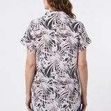 Back view of a woman wearing a short rolled-sleeve button-front blouse in black and blush palm leaf print, paired with blue