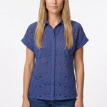 Blonde female model wearing a blue eyelet button-front short-sleeve blouse with floral cutwork detail, paired with dark