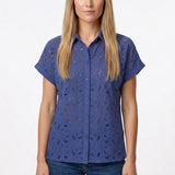Blonde female model wearing a blue eyelet button-front short-sleeve blouse with floral cutwork detail, paired with dark