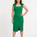 Brunette woman wearing an emerald green sleeveless sheath dress with square neck, waist band, front pleating, and front