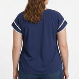 Plus-size model shows back view of navy blue short-sleeve popover blouse with white pom-pom trim on flutter sleeves, paired