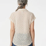 Back view of a woman with gray hair wearing a blush floral lace collared blouse with cuffed cap sleeves over blue jeans.