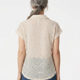 Back view of a woman with gray hair wearing a blush floral lace collared blouse with cuffed cap sleeves over blue jeans.