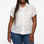 Plus-size model wears a white short-sleeve eyelet button-front blouse with floral cutwork detail; front view, waist up.